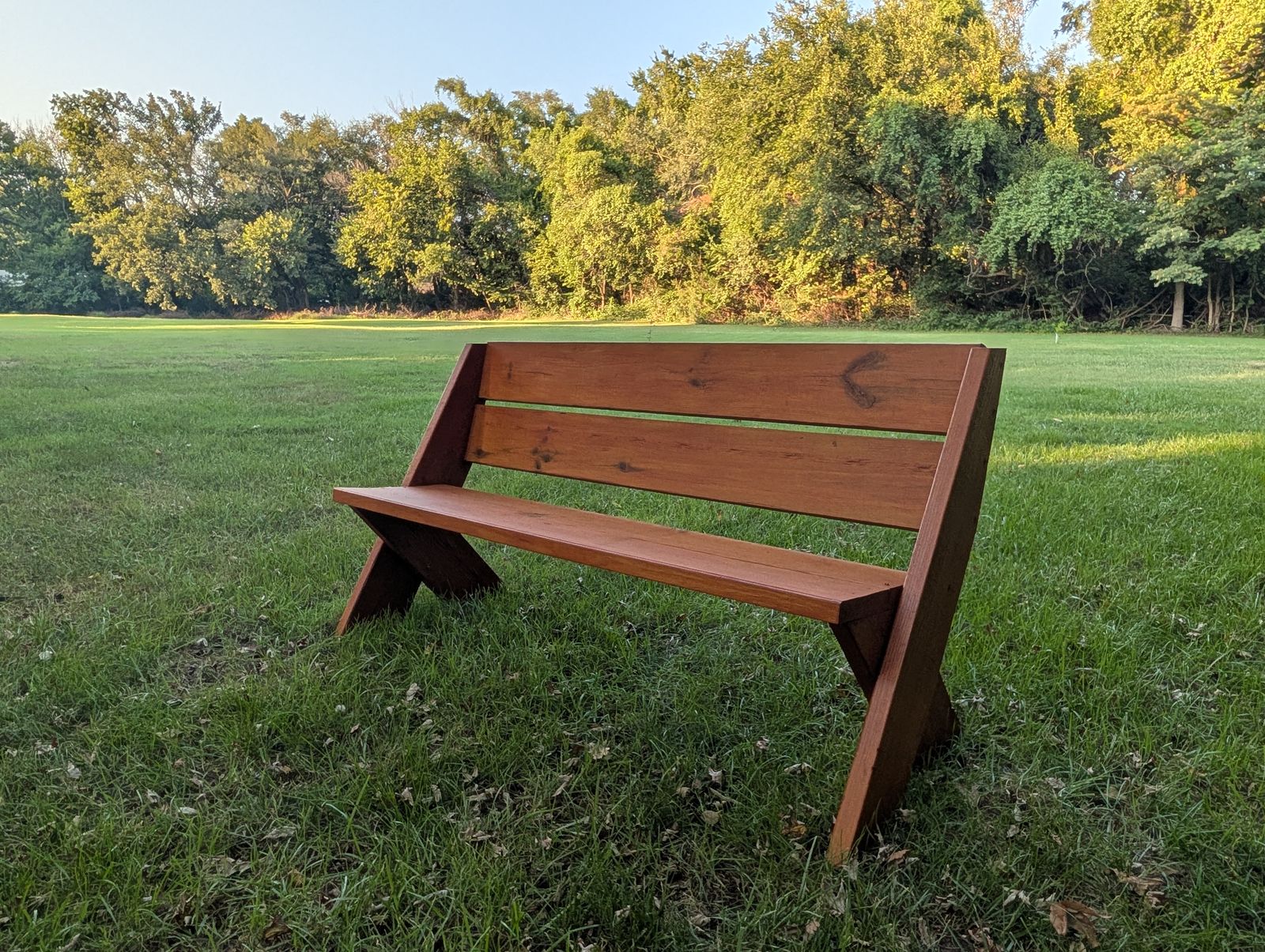 A Leopold bench in solid pine with a warm finish, standing in a sunlit field at the edge of the woods